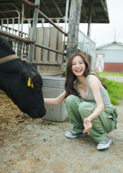 Ms. Mariho Kokei is a Japanese & Asian fashion model, swimsuit model (gravure idol / bikini model / pin-up girl), actress, and former idol, she's wearing a gray running shirt and green pants, she's on a farm, beside a black cow, she's crouching.