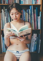 Ms. Ami Ojimi is a young gravure idol (bikini model / swimwear model) and a young and cute actress, she is wearing a white bikini, sitting on the floor, reading a book, and behind her is a bookshelf.