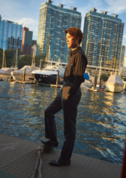Mr. Ayato Akaibashi is standing next to several boats, wearing a dark cut shirt and pants, he is also an actor and voice actor.