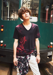 Mr. Ayato Akaibashi is standing in the back of a green bus wearing a red short-sleeved shirt and white pants, he is also an actor and voice actor.
