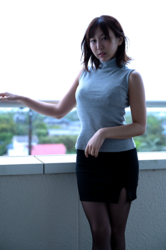 Ms. Kirina Hinaga is wearing a gray shirt and a black skirt and she is standing on the corridor in the apartment, she is a Japanese & Asian sweet and cute big breasts gravure idol (pin-up model, bikini model, swimsuit model), her bust is 90 cm, she has charming big breasts, she is a woman with sexual charm.