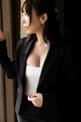 Ms. Kirina Hinaga is wearing a black women's suit and white shirt and is standing in a room, she is a Japanese & Asian sweet and cute big breasts gravure idol (pin-up model, bikini model, swimsuit model), her bust is 90 cm, she has charming big breasts, she is a woman with sexual charm.