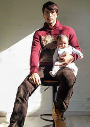 Mr. Rodrigue Takise is wearing red clothes, brown pants, and is sitting on a chair with a baby on his left thigh, he is a half Japanese half French handsome mixed-race male model.