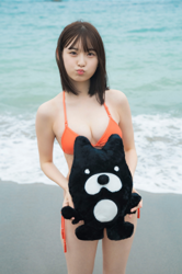 Ms. Asumi Saigosawa is wearing an orange bikini swimsuit and is standing on the beach holding a stuffed black bear in her hand, she is a very cute and young actress who is also active as a bikini model (swimsuit model / gravure idol / pin up girl).